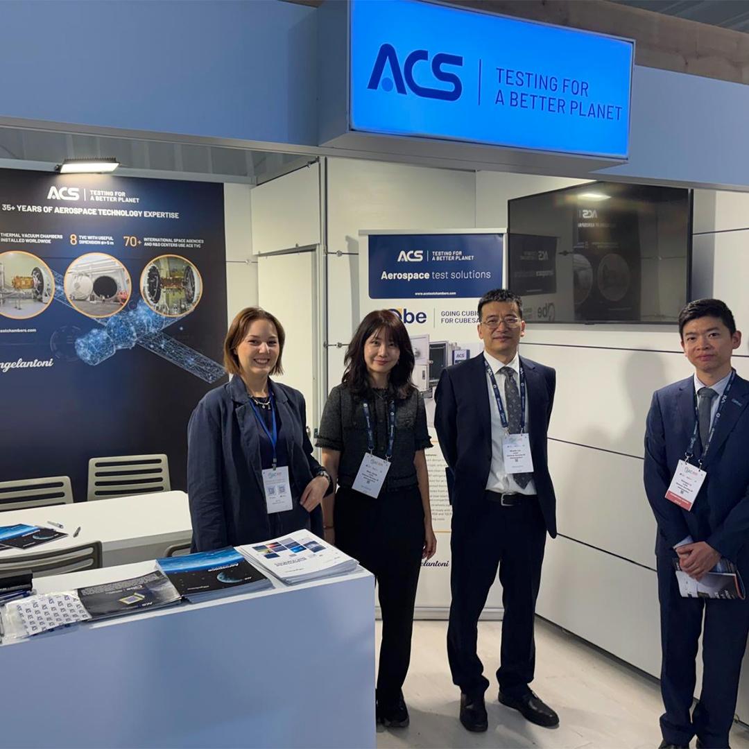 Des membres de l'équipe d'Angelantoni Test Solutions (ACS) sur le stand d'exposition lors de l'International Astronautical Congress (IAC) avec des bannières et des brochures sur les tests aérospatiaux.